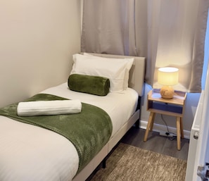 A neatly made bed with a green throw, a white towel, and a green pillow. There is a bedside table with a lamp and a small wooden stool. - St Julian House- Town Centre- Castle Park- Hot Tub (Colchester)
