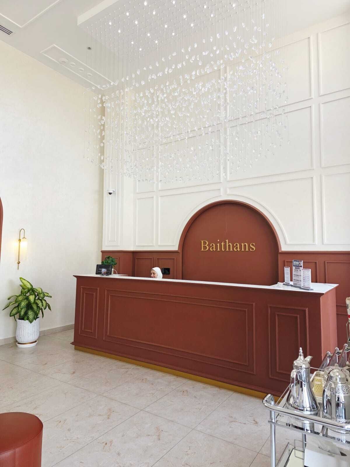 A reception desk with a nameplate reading 'Baithans' in a hotel lobby with a chandelier, a potted plant, and a coffee service cart.