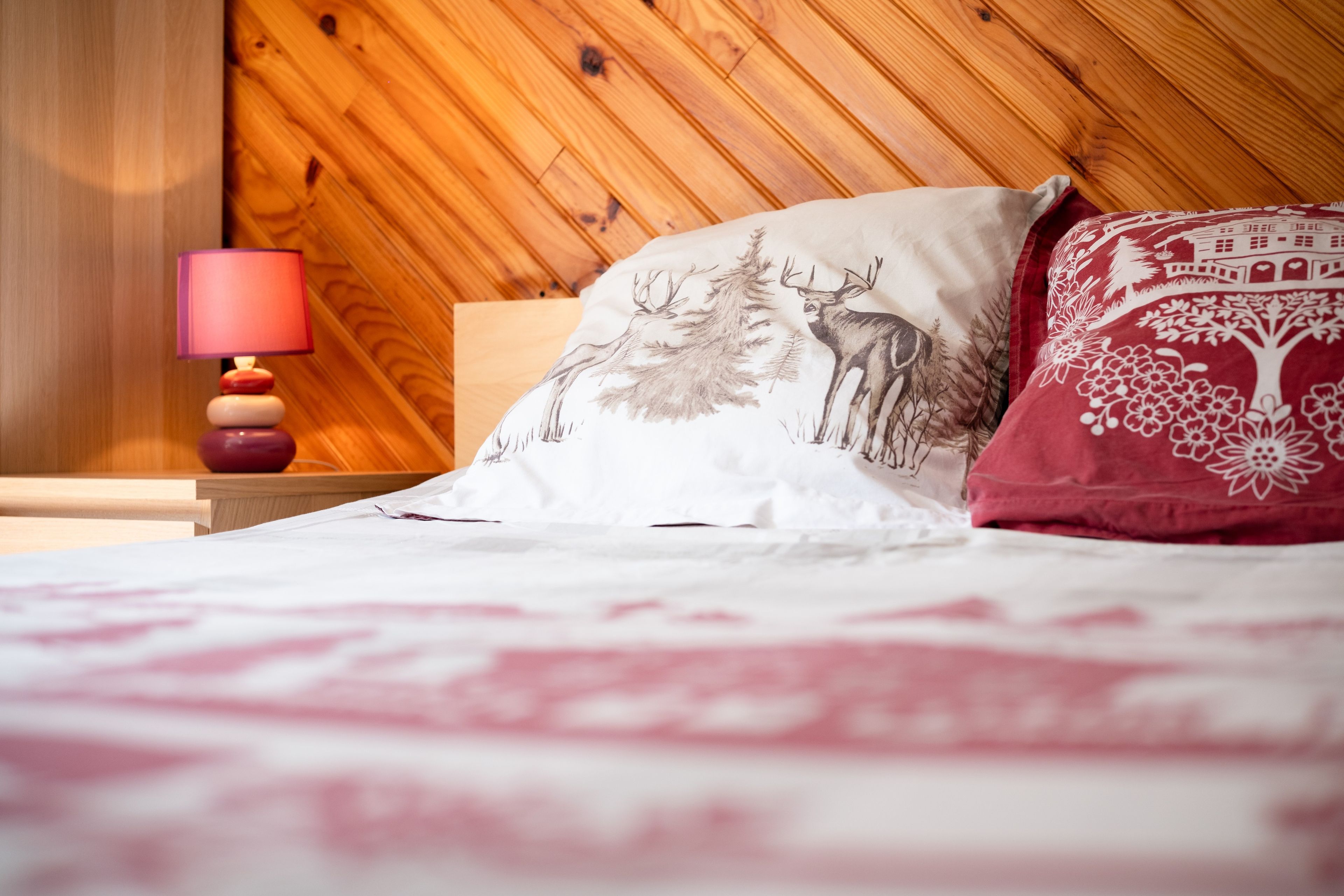 Een bed met een deken met een wit en rood patroon, een wit kussen met een ontwerp van een bos en herten, en een rood kussen met een bloemenmotief.