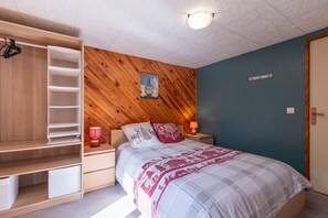 A bedroom with a bed, a nightstand, a wooden wardrobe, and a wall-mounted shelf.