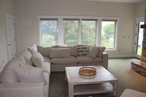 A living room with a beige sectional sofa, a small white coffee table, and a large window with a view of greenery. - Vista Cove Lake Lure- Private Cove & Game Room (Lake Lure)
