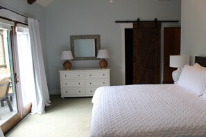 Quarto com cama, cômoda com duas luminárias, espelho e porta para outro aposento.