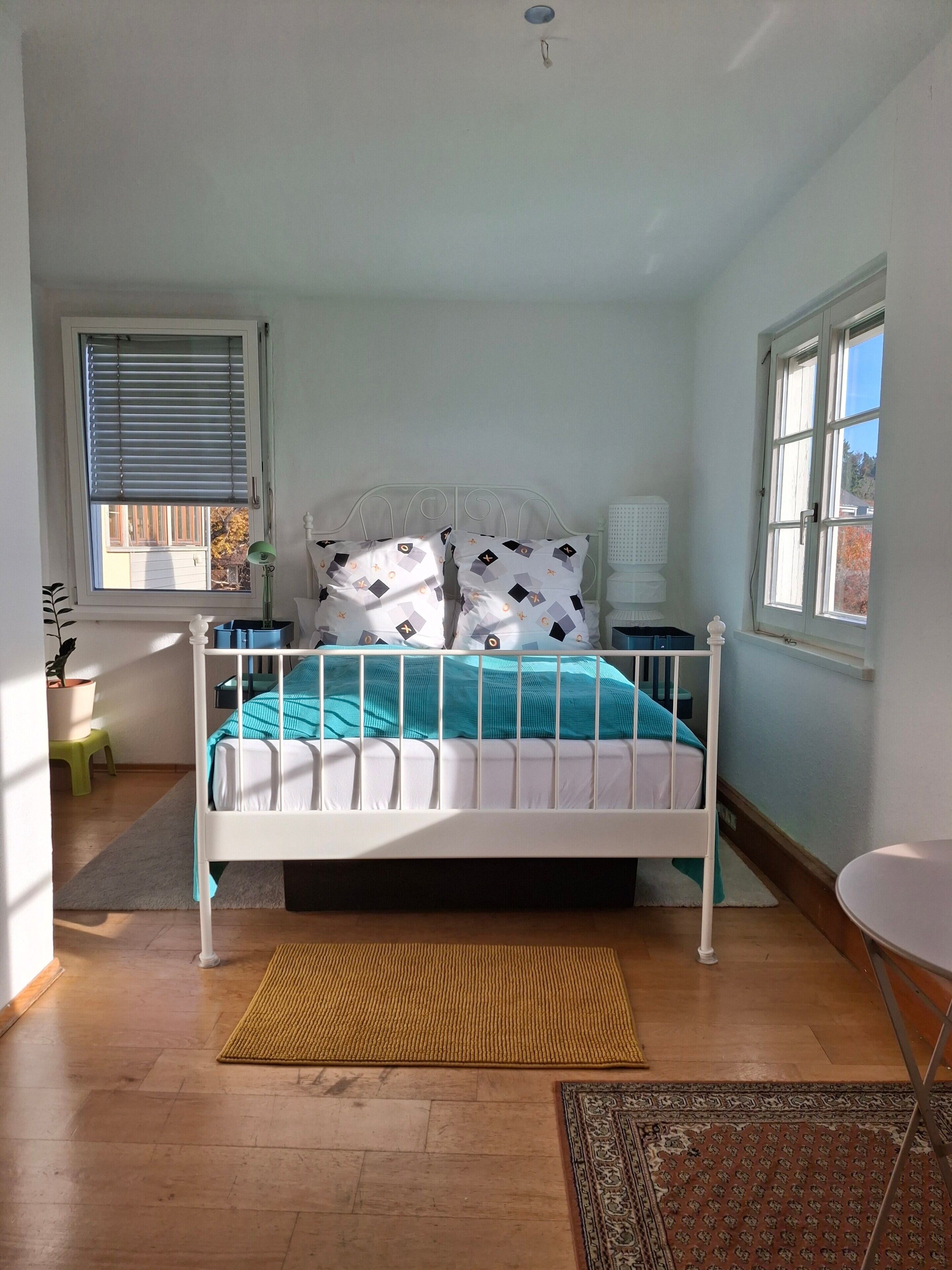Ein Schlafzimmer mit einem Bettgestell aus weißem Metall, einer türkisblauen Tagesdecke und gemusterten Kissen. Es gibt ein Fenster mit Jalousien, einen kleinen Hocker und eine Lampe auf einem Nachttisch.