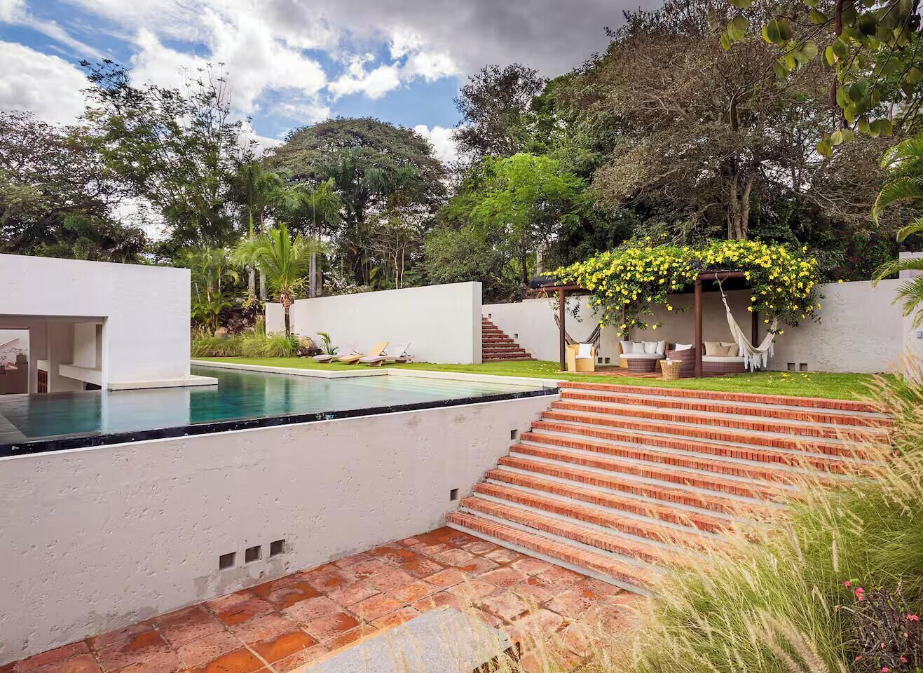 Ein moderner Poolbereich im Freien mit freiem Blick zum Himmel, umgeben von üppigem Grün und einer Treppe zu einer überdachten Sitzgelegenheit.