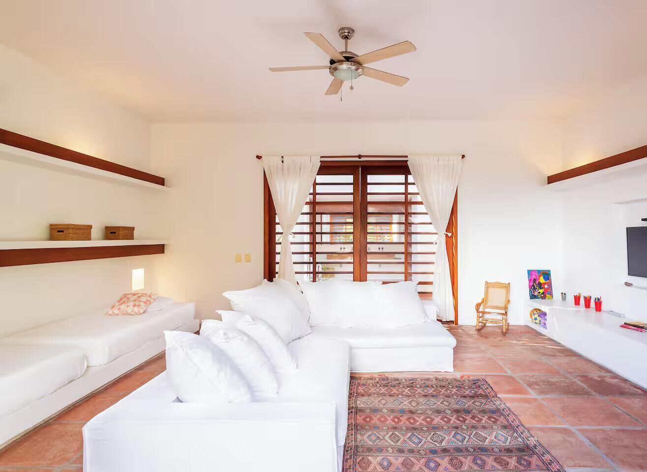 A modern living room with a white sofa set, a ceiling fan, a patterned rug, and a TV on the wall.