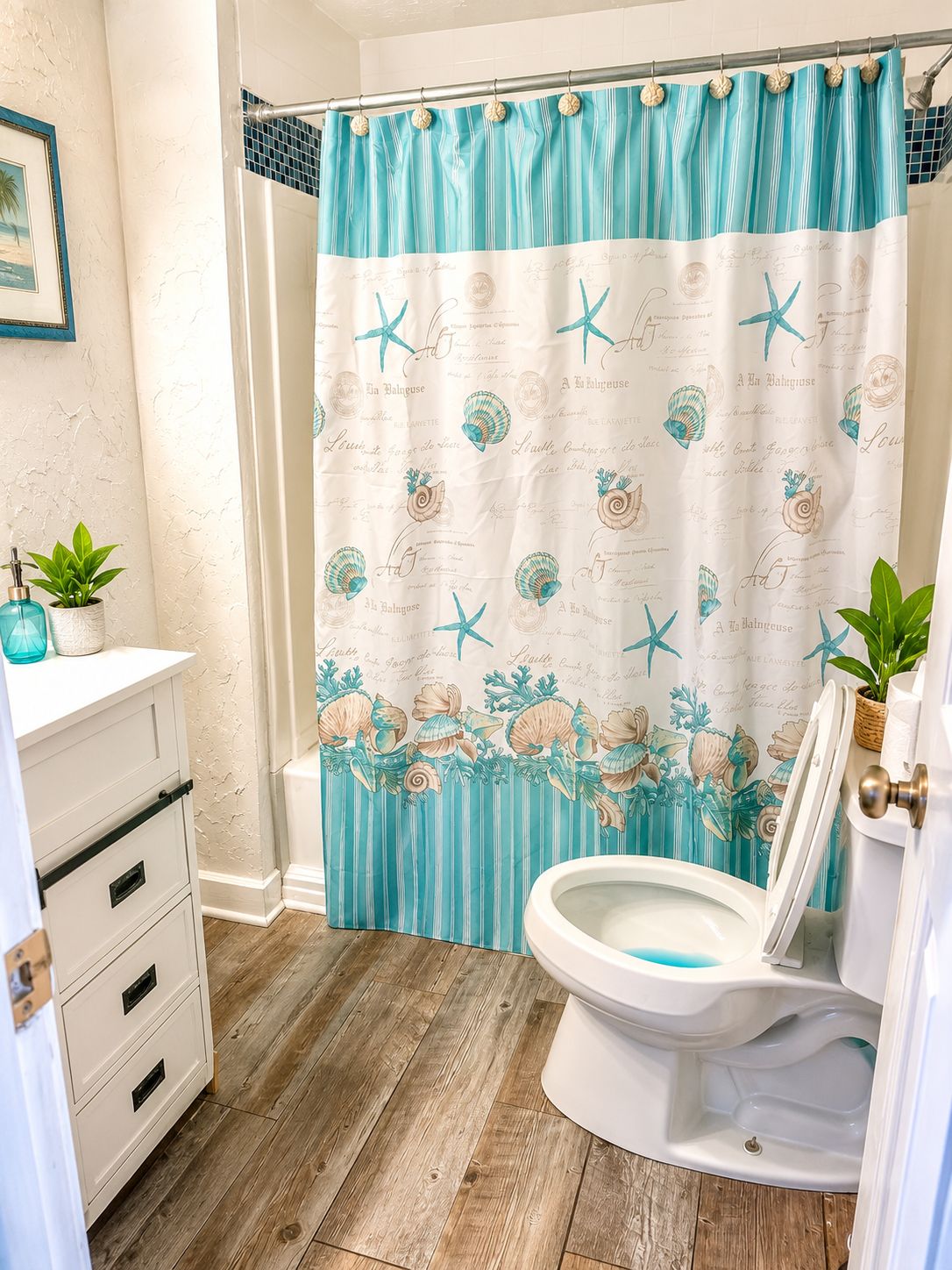 A bathroom with a wooden floor, a white toilet, a shower curtain with sea-themed designs, and a small potted plant on the counter.