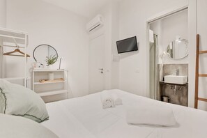 Habitación de hotel con cama, televisor montado en la pared, baño con lavamanos y espejo, y repisa con artículos de aseo.