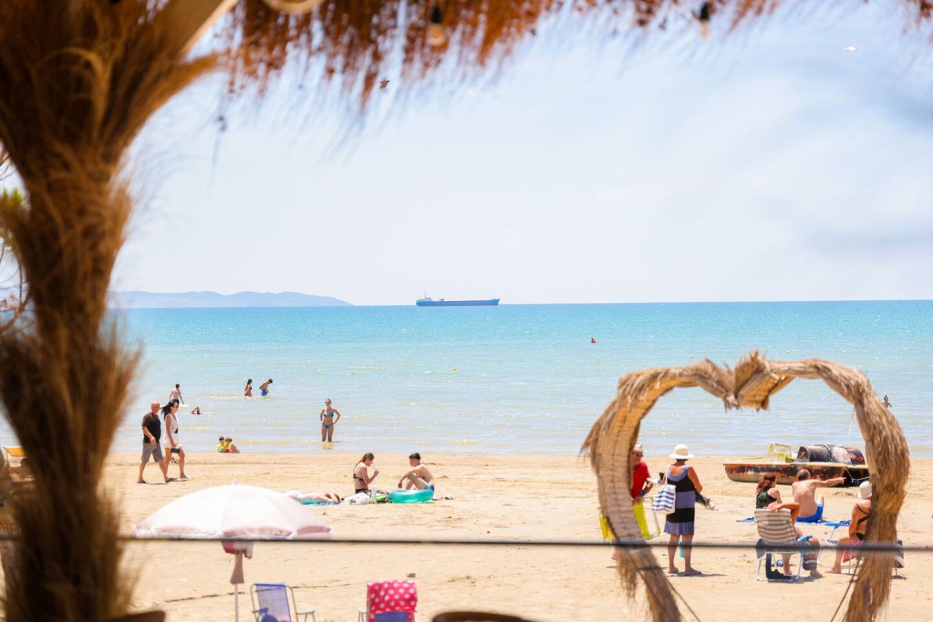 A beach scene with a large ship in the distance, people sunbathing, and a makeshift arch structure on the sand.