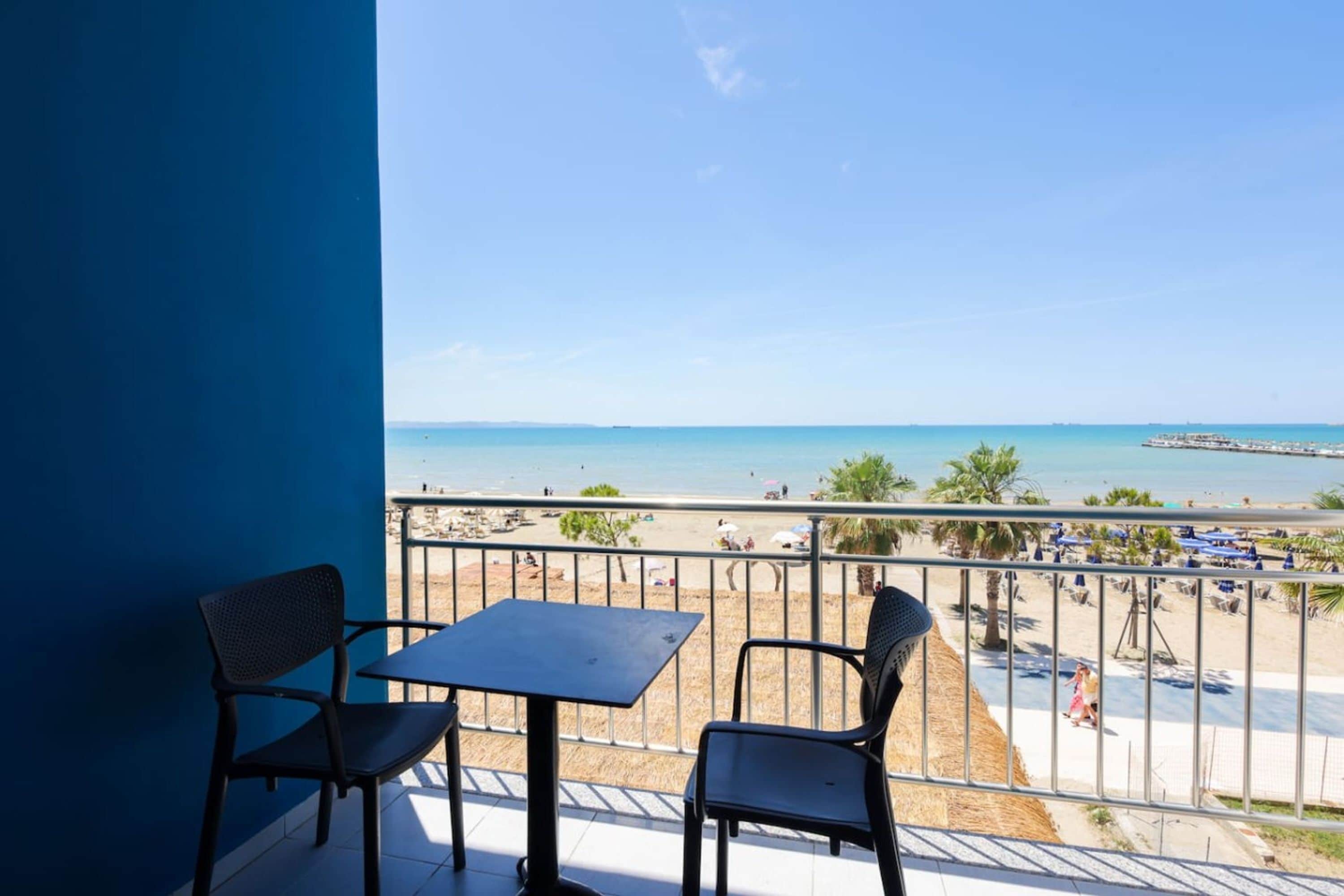 Een balkon met twee stoelen en een tafel, met uitzicht op een strand met parasols en een pier.
