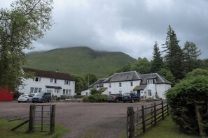 A rural setting with white buildings, a gravel driveway, and a mountain in the background. - Curlew Cottage (Crianlarich)