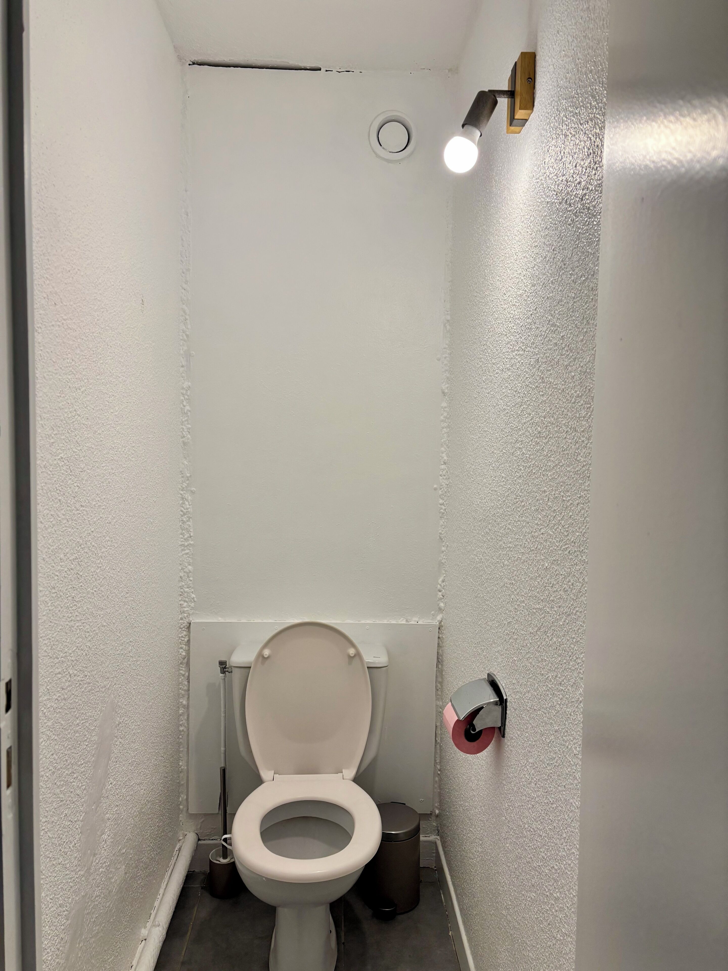 Une petite salle de bain aux murs blancs, équipée de toilettes, d’une poubelle et d’un luminaire.