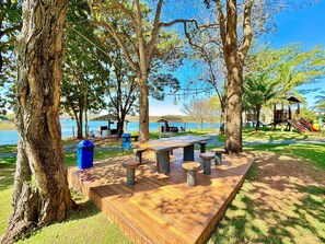 A picnic area with a wooden table and benches, surrounded by trees and a view of a body of water. - Pontal do Lago Flat Residence, Aproveite Dias Incriveis em Familia as Margens do Lago Corumba (Caldas Novas)