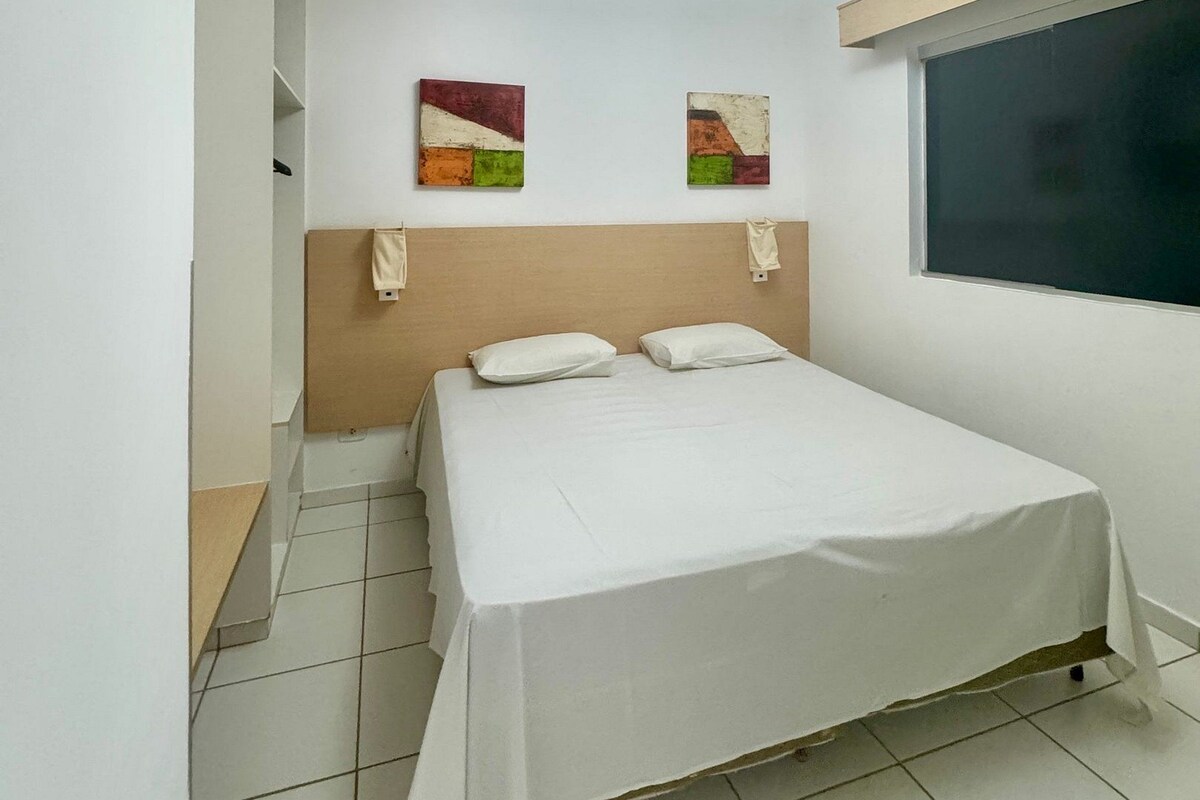 A single-bed room with a white bedspread, two wall-mounted paintings, a window with a dark blind, and a white ceiling light.