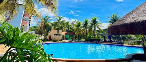A clear blue pool surrounded by palm trees and a thatched-roof structure, with a view of a modern building in the background.