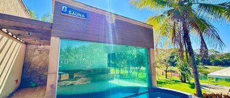 A modern spa building with a glass enclosure, a sign reading 'SAUNA', and a pool area with a clear view of the surrounding greenery.