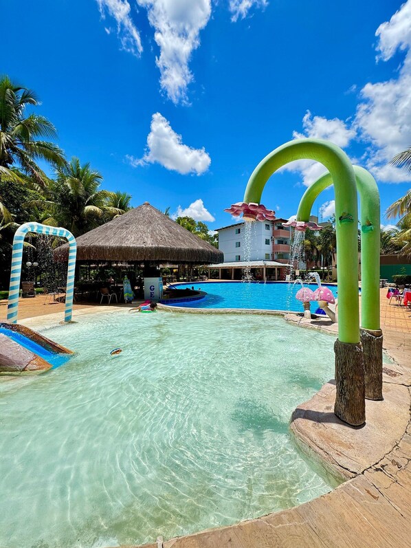 A swimming pool with a slide and a thatched-roof structure, surrounded by palm trees and a clear blue sky. - Pontal do Lago Flat Residence, Aproveite Dias Incriveis em Familia as Margens do Lago Corumba (Caldas Novas)