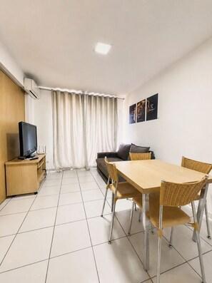 A room with a TV on a wooden stand, a sofa, a dining table with chairs, and a window with curtains. - Pontal do Lago Flat Residence, Aproveite Dias Incriveis em Familia as Margens do Lago Corumba (Caldas Novas)
