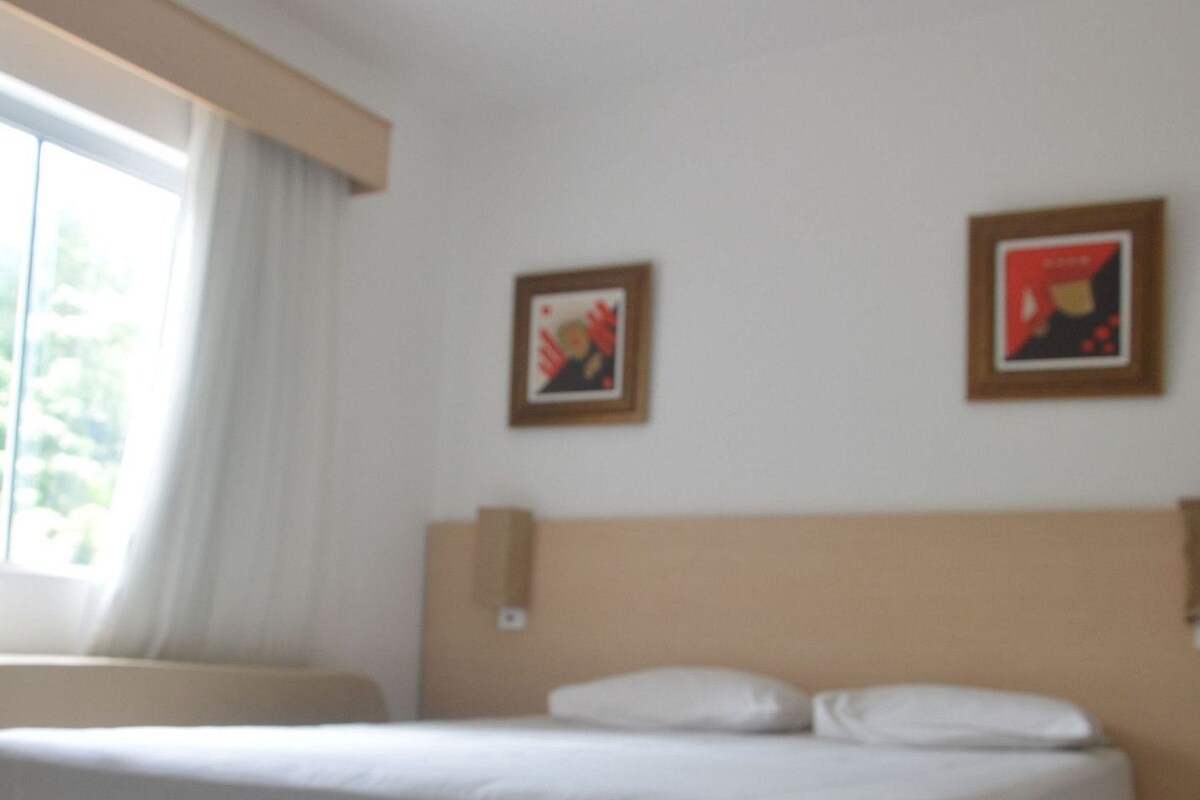 A hotel room with a bed, a window with curtains, and two framed pictures on the wall.