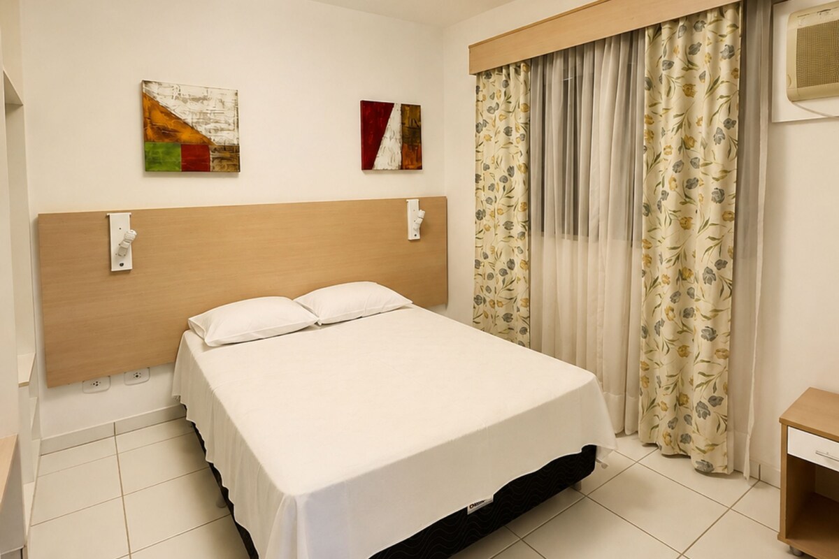 A hotel room with a bed, a headboard, a wall-mounted light fixture, a flat-screen TV, and a window with curtains.