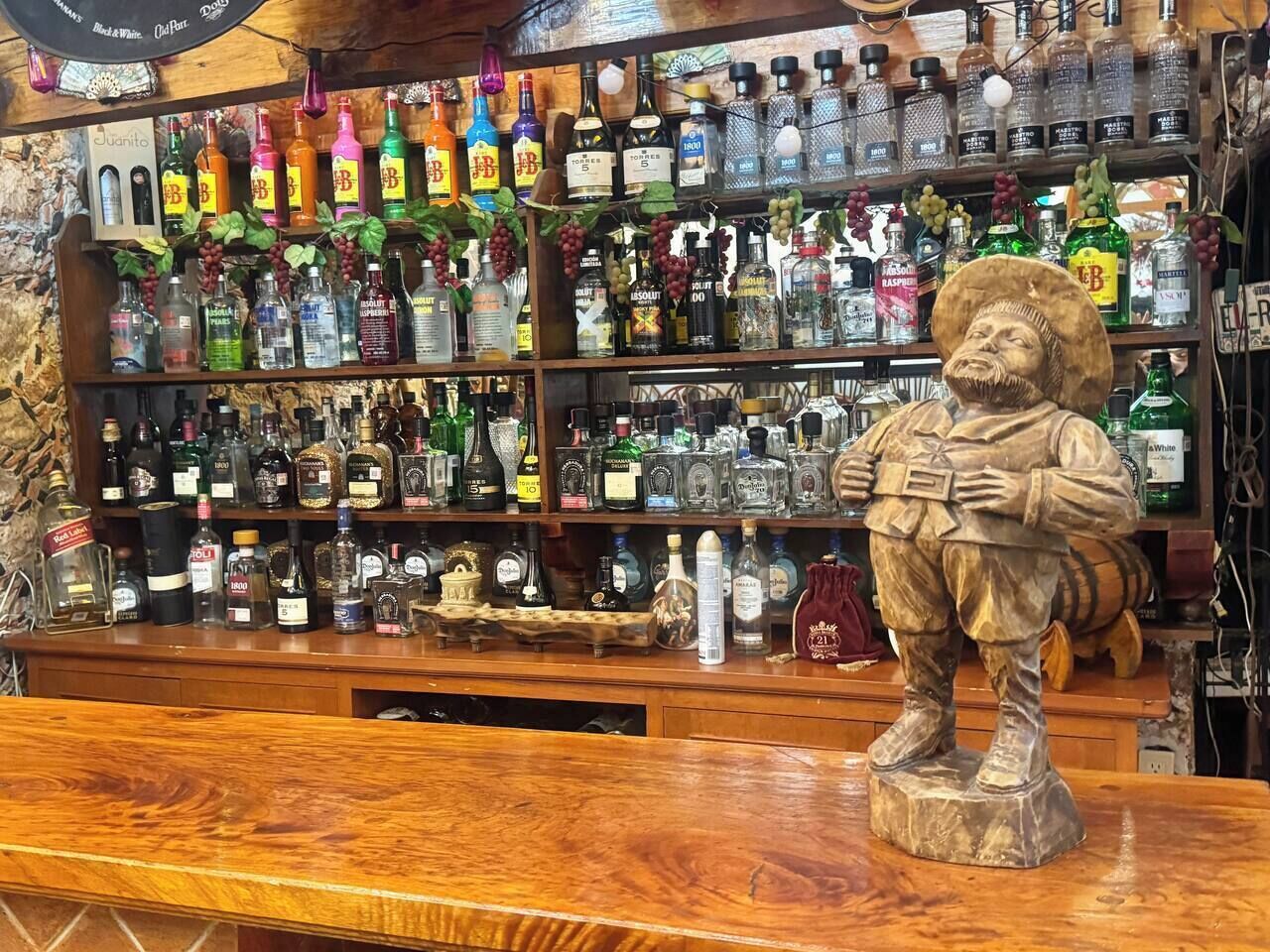 En bar med en trædisk og en statue af en person, der holder en flaske. Bag disken er der hylder fyldt med diverse flasker med spiritus.