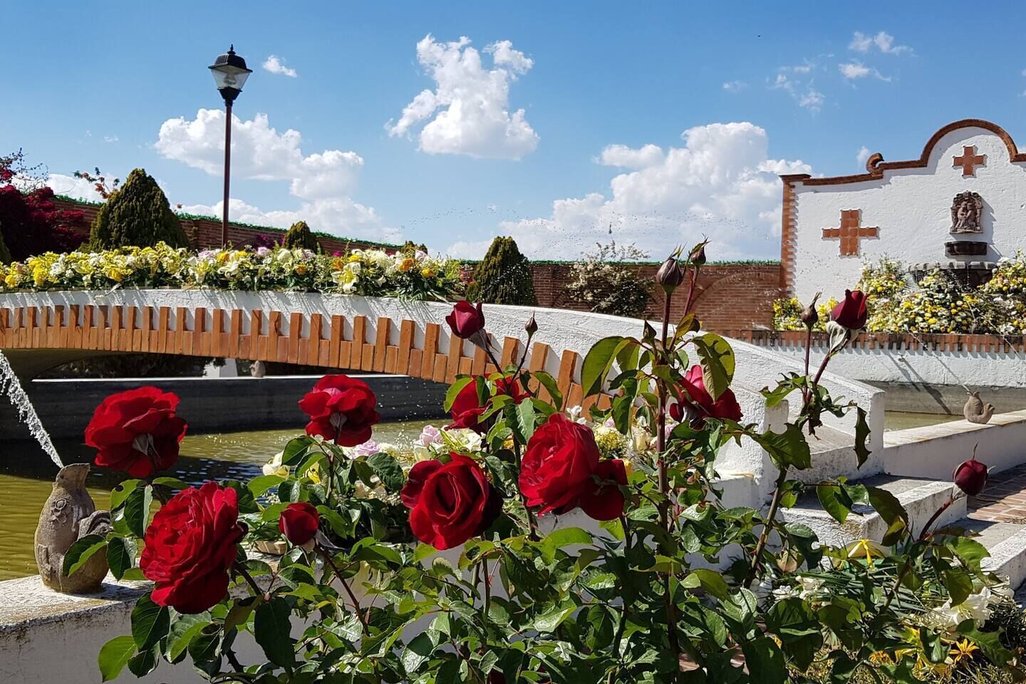 Un jardin avec un pont, un bâtiment blanc surmonté d’une croix et une statue.