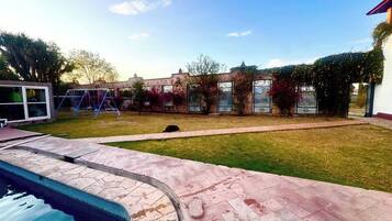 A swimming pool with a stone deck, a grassy lawn, and a building with a brick facade in the background.