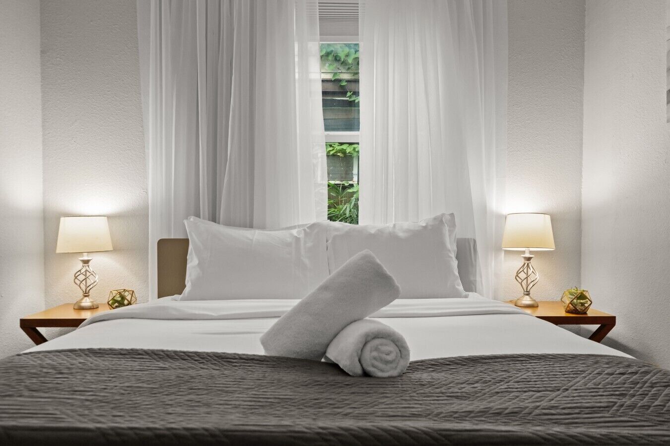 A neatly made bed with white linens and a rolled towel, two bedside lamps, and sheer curtains.