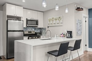 Une cuisine moderne avec des meubles blancs, des appareils en acier inoxydable, un îlot central doté de tabourets de bar, un éclairage suspendu et des œuvres d’art accrochées aux murs.
