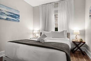 Une chambre d’hôtel avec un lit, deux tables de chevet, une fenêtre avec des rideaux blancs et un tableau au mur.