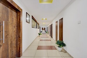 A hotel corridor with wooden doors, framed artwork, potted plants, and a tiled floor.