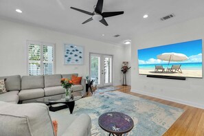 A living room with a sofa, coffee table, and a large TV screen displaying a beach scene. - Sunset Serenity-Minutes away from the beaches, Old Town and the best of KW! (Key West)