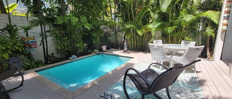 Un espace piscine agrémenté d’une terrasse en bois, de meubles en osier blancs et d’une végétation tropicale luxuriante.