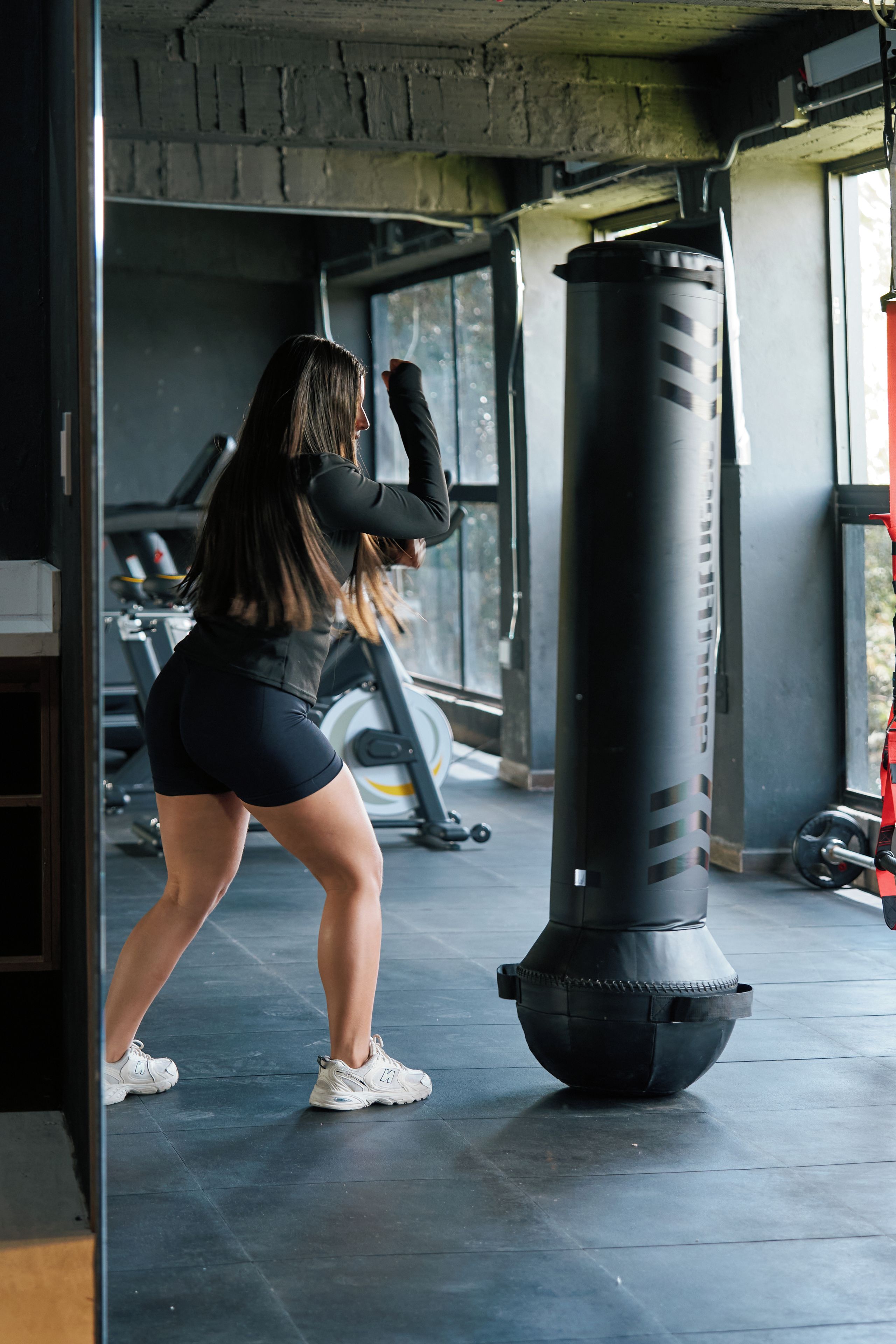 Een vrouw die aan het trainen is met een bokszak in een sportschool, met fitnessapparatuur op de achtergrond.