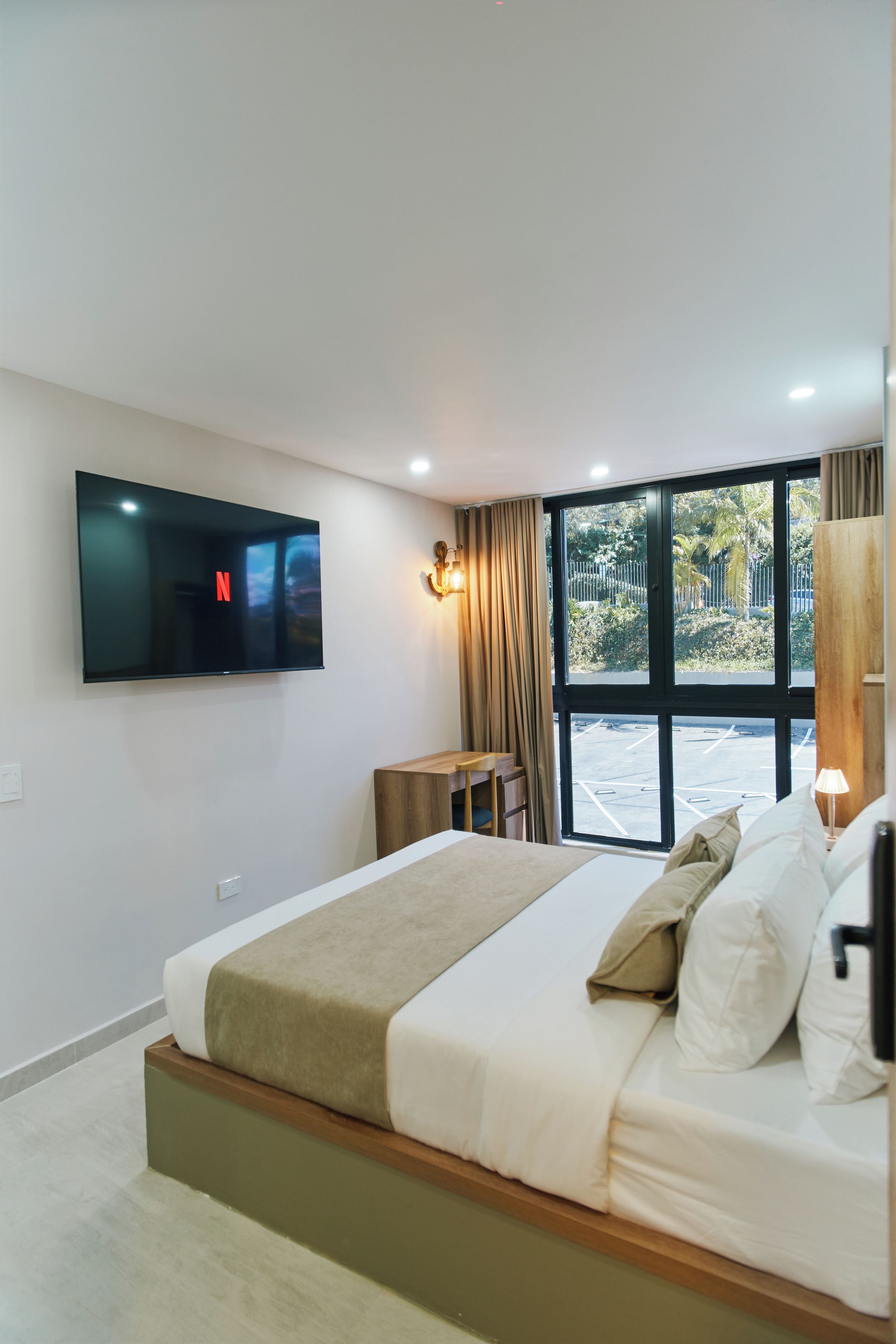 A modern hotel room with a large bed, a flat-screen TV mounted on the wall, a desk with a chair, and a large window offering a view of the outdoors.