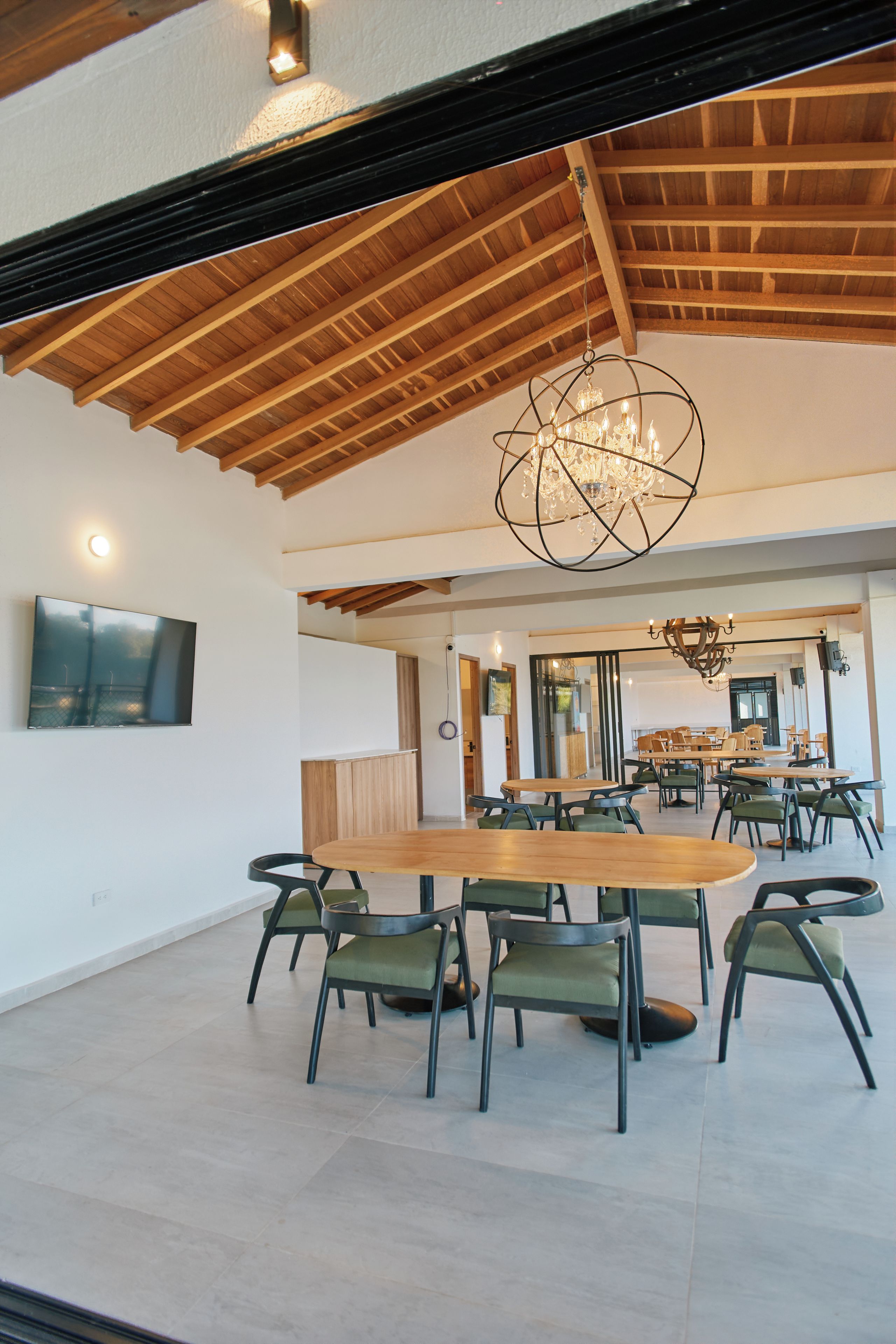 A spacious dining area with wooden tables and chairs, a chandelier, and a television mounted on the wall.