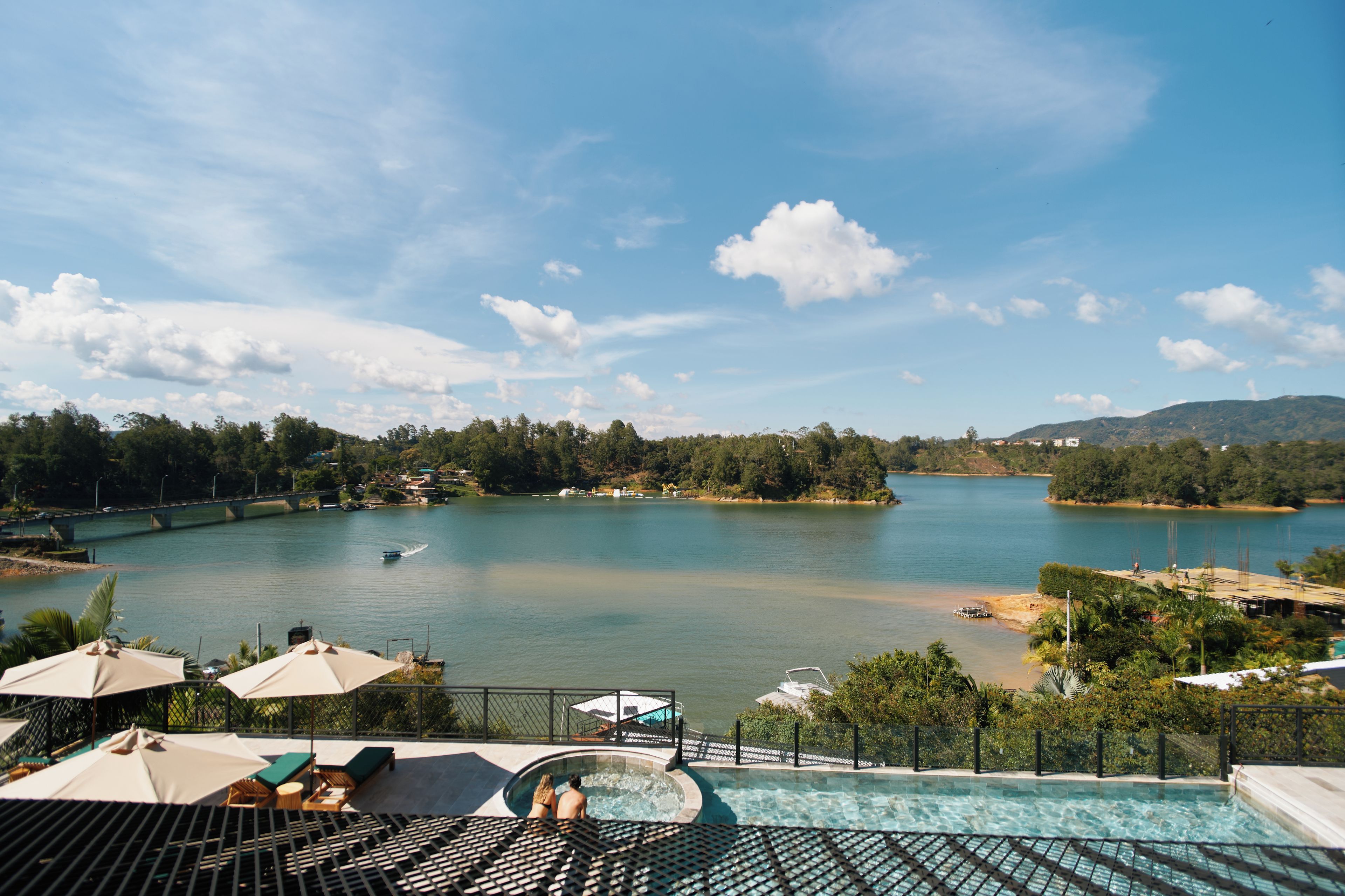 A pool area with lounge chairs and umbrellas, overlooking a calm lake with a bridge and buildings in the background.