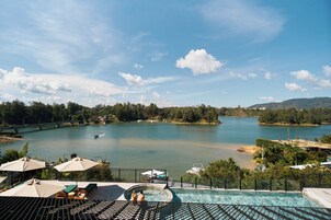 Zona de piscina con sillones de descanso y sombrillas, con vista a un lago tranquilo con un puente y edificios al fondo.
