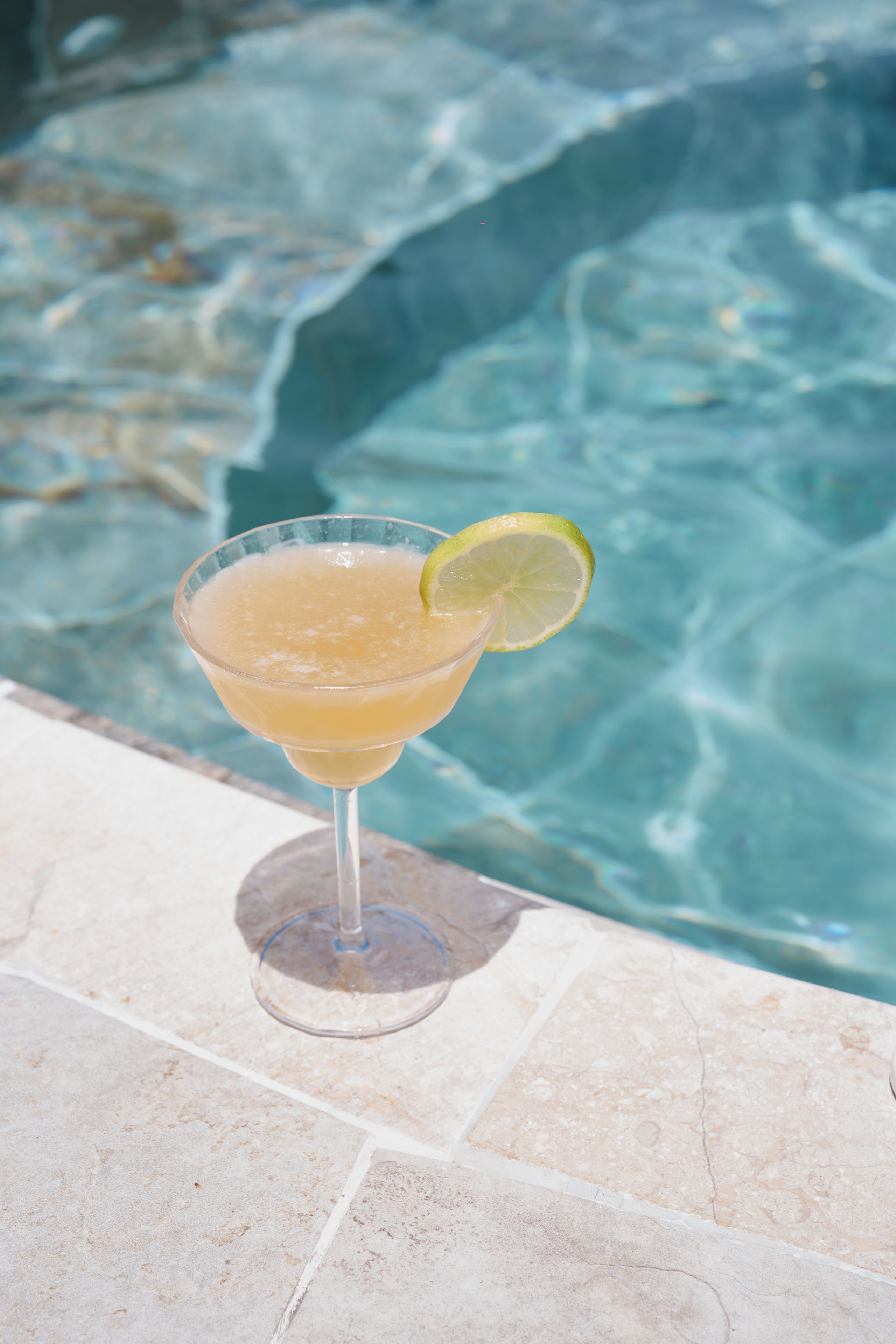Un verre à cocktail, décoré d’une tranche de citron vert, posé sur une table carrelée au bord de la piscine.