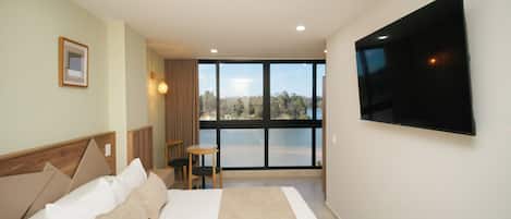 A hotel room with a bed, a TV mounted on the wall, a window with a view of the outdoors, and a small table with chairs.