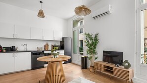 A modern kitchen with white cabinets, a dining area with a round table, and a living area with a TV and a plant. - Le Foch - Bel appt moderne à 20 min de la plage (Béziers)