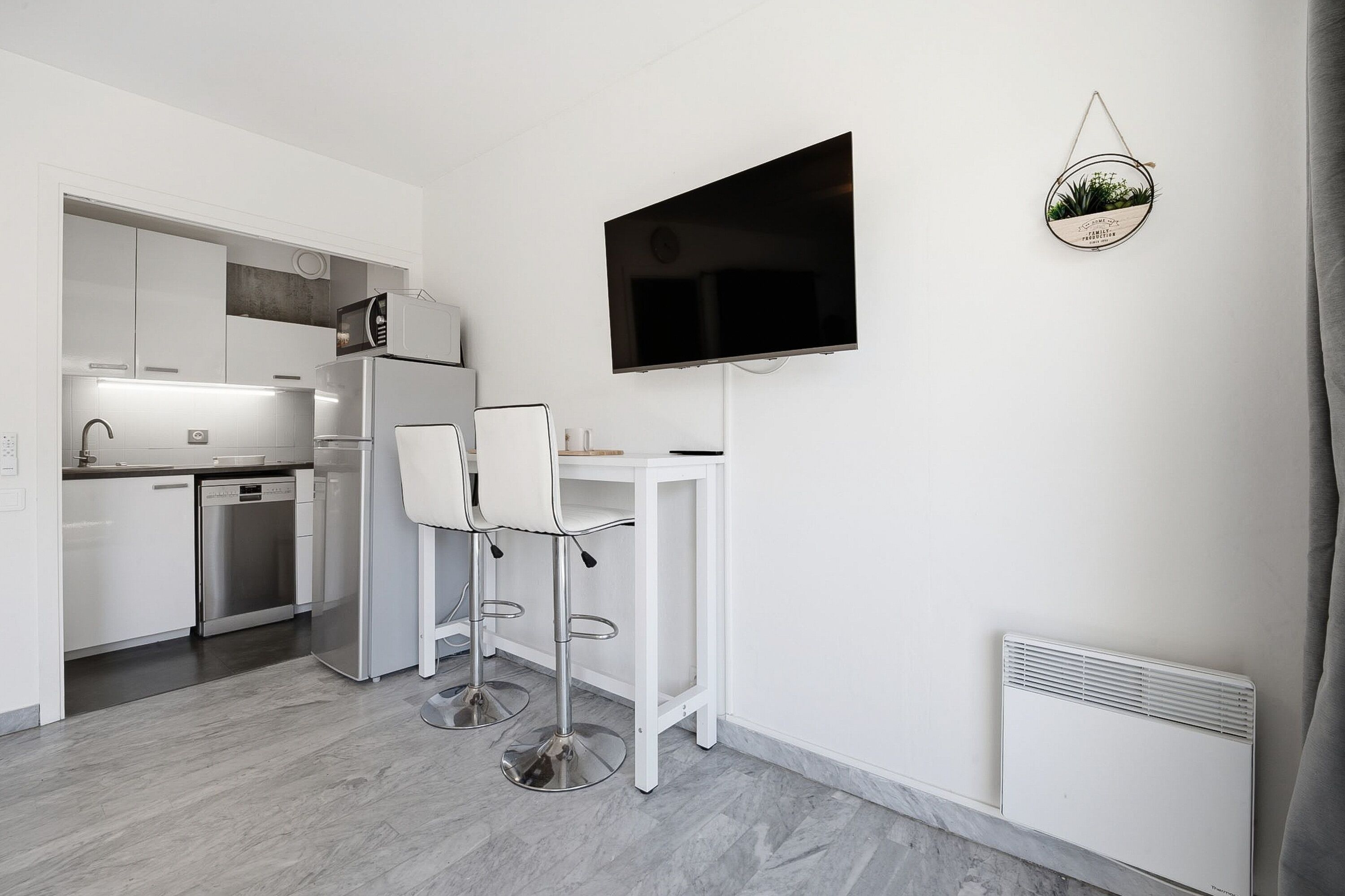 A modern kitchen with white cabinets, a built-in microwave, and a refrigerator. There are two white bar stools and a flat-screen TV mounted on the wall.