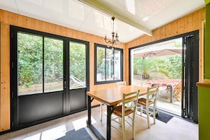 A dining area with a wooden table and chairs, large windows, and a view of the outdoors. - Les Muguets - Maison à 5 min de la plage (Sainte Brevin-les-Pins)