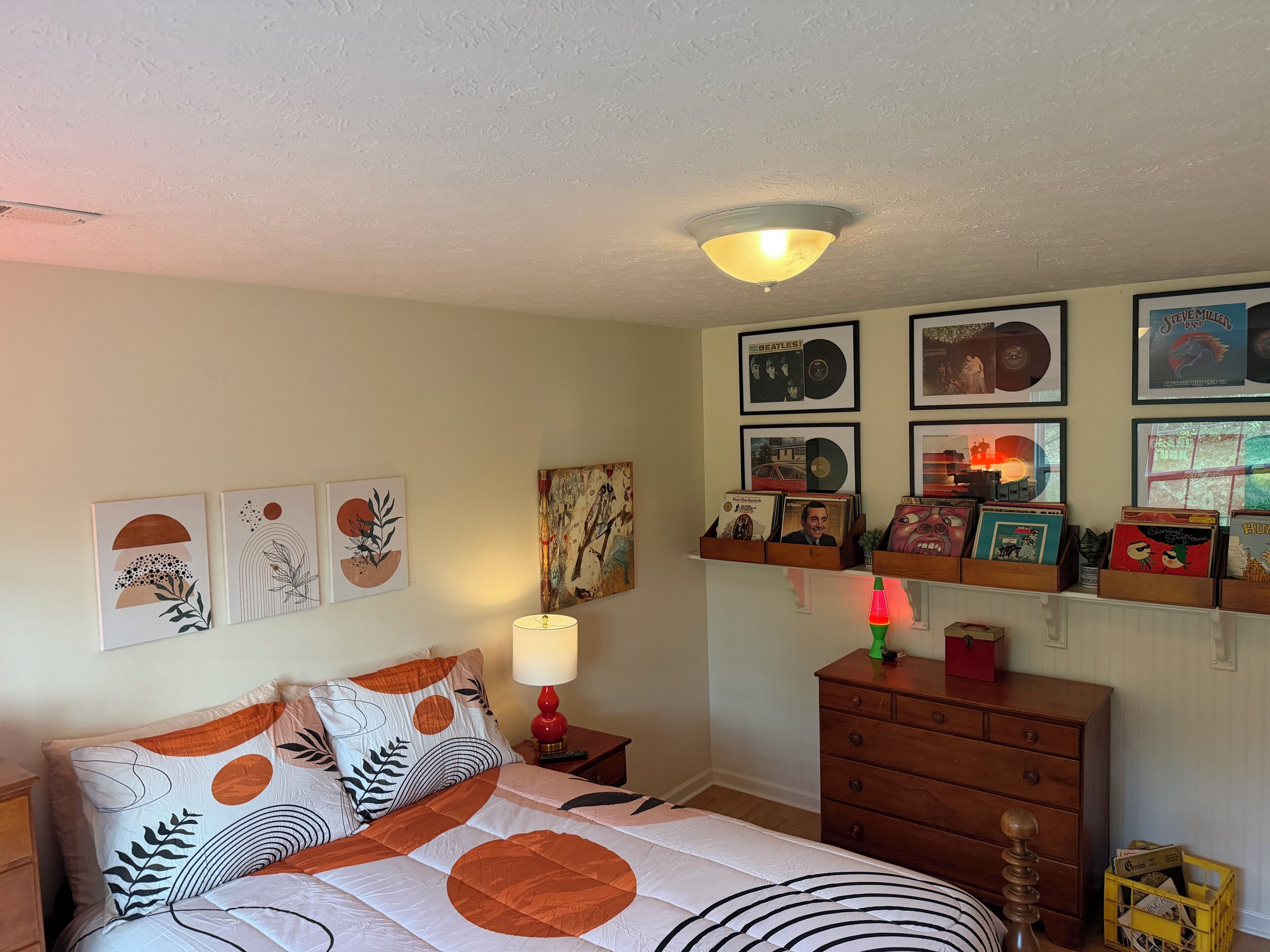 A bedroom with a bed, a dresser, a nightstand, a lamp, and wall-mounted shelves with framed items.