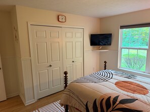 Een slaapkamer met een bed, een televisie op een plank, een deur en een raam met uitzicht op groen.