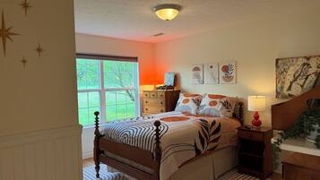 A bedroom with a large window, a bed with patterned bedding, a dresser, and wall art.