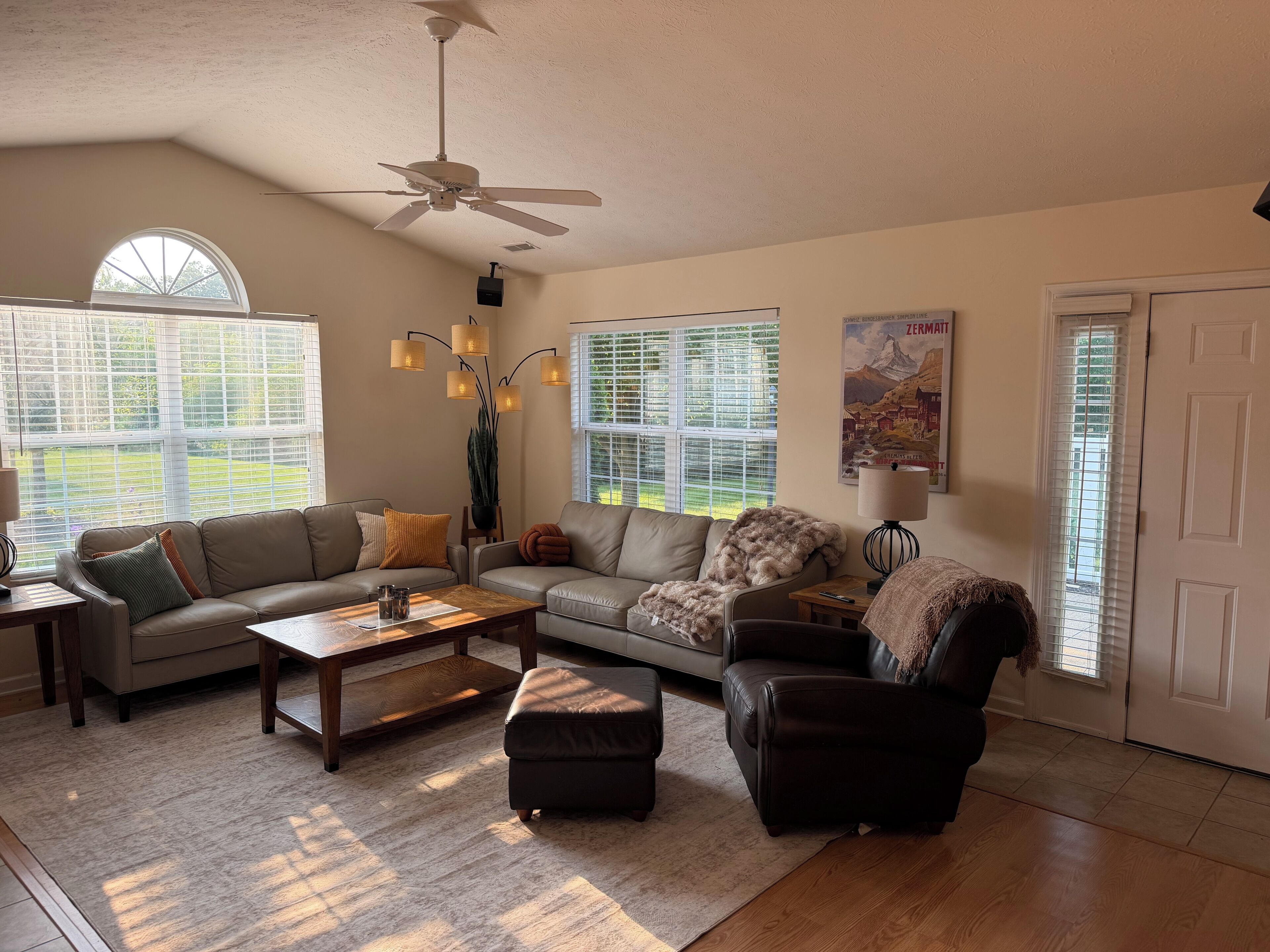 A living room with a ceiling fan, large windows, a sofa set, a coffee table, and a lamp.