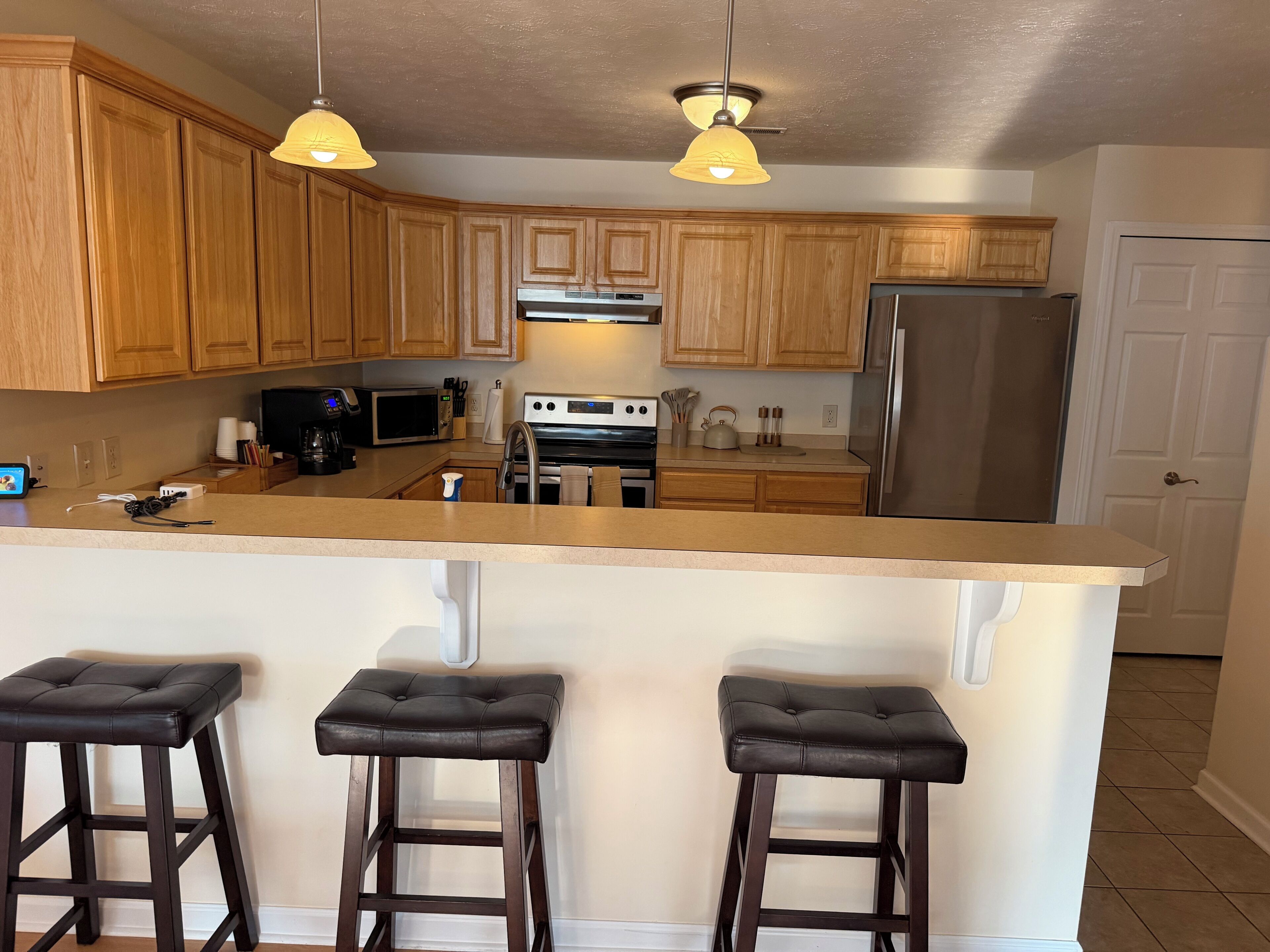A kitchen with wooden cabinets, a stainless steel refrigerator, a microwave, and a stove. There are three bar stools and pendant lights above the counter.