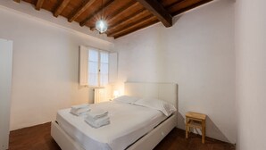 Ein Schlafzimmer mit einem Einzelbett, einem kleinen Holzschemel, einem Nachttisch mit Lampe und einem Fenster mit Vorhängen.