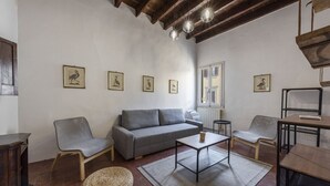 Ein Wohnzimmer mit grauem Sofa, zwei Sesseln, einem Couchtisch und einem gewebten Hocker. An der Wand hängen gerahmte Bilder, ein Fenster mit Fensterläden und eine Holzbalkendecke.