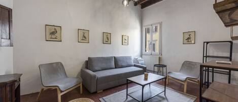 Un salón con un sofá gris, dos sillones, una mesa de centro y un taburete tejido. Hay cuadros enmarcados en la pared, una ventana con contraventanas y un techo de madera.